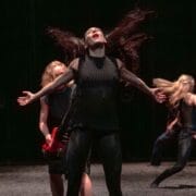 Three dancers in dark clothing perform on stage; the central dancer faces forward with hair flying and arms outstretched, while the others move dynamically in the background.