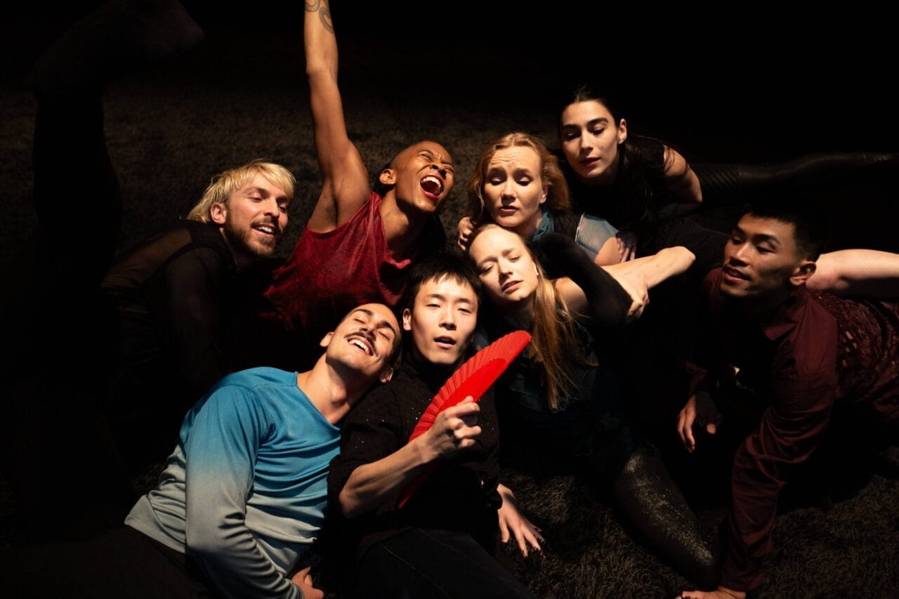Eight people are closely grouped together on a dark stage, some with eyes closed and others smiling or laughing; one person in front holds a red fan.