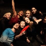 Eight people are closely grouped together on a dark stage, some with eyes closed and others smiling or laughing; one person in front holds a red fan.