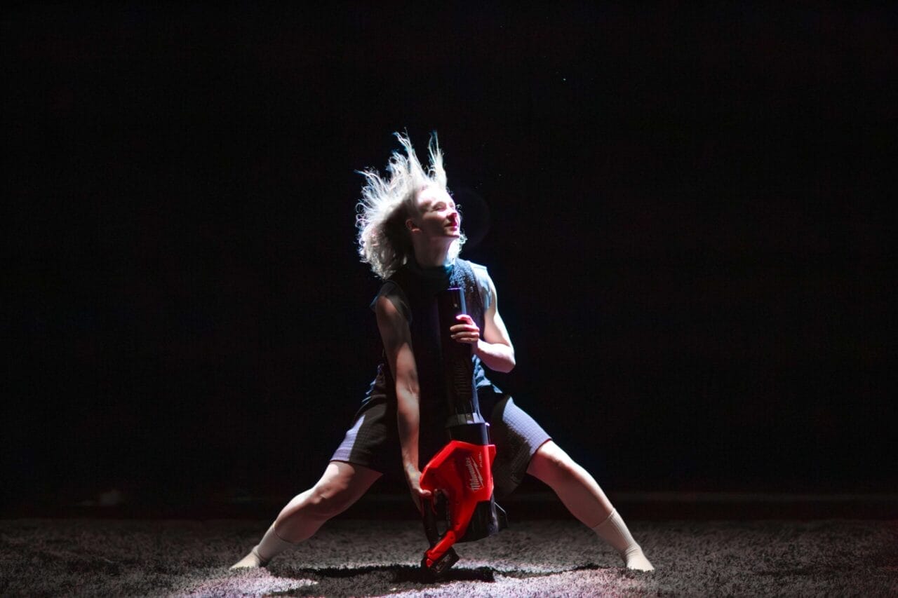 A person with light hair and a hat stands under a spotlight, holding a red leaf blower, with legs bent wide and hair flying upward.