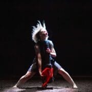 A person with light hair and a hat stands under a spotlight, holding a red leaf blower, with legs bent wide and hair flying upward.