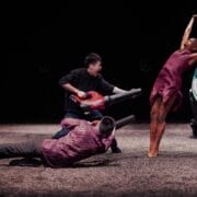 Three performers are on stage; one woman in a dress poses with arms raised, while two men point leaf blowers at her. Another person stands in the background.