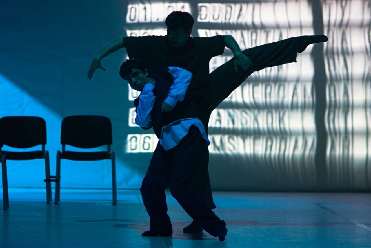 Two dancers perform on stage; one lifts the other while extending a leg sideways. The scene is lit with blue light, and two chairs and a projected background are visible.