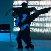 Two dancers perform on stage; one lifts the other while extending a leg sideways. The scene is lit with blue light, and two chairs and a projected background are visible.