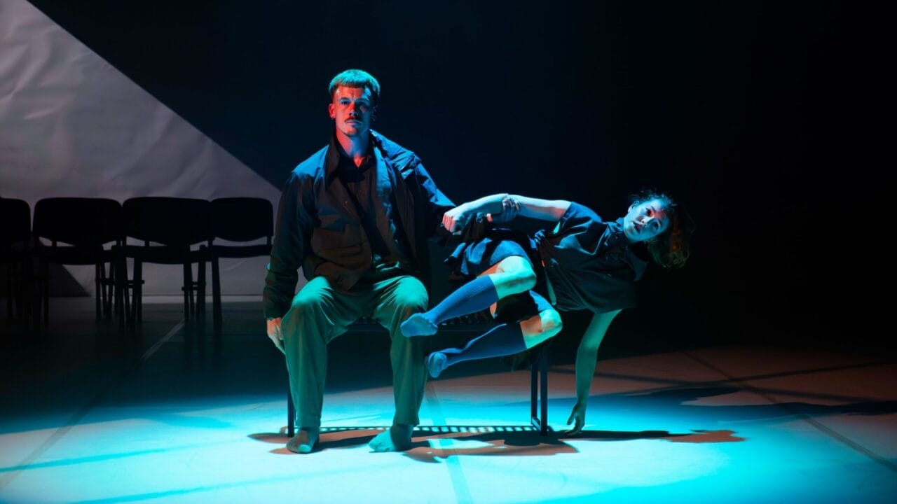 A man sits on a bench under stage lighting while a woman reclines across his lap, both barefoot and dressed in dark clothing on a dimly lit stage.