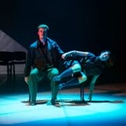 A man sits on a bench under stage lighting while a woman reclines across his lap, both barefoot and dressed in dark clothing on a dimly lit stage.