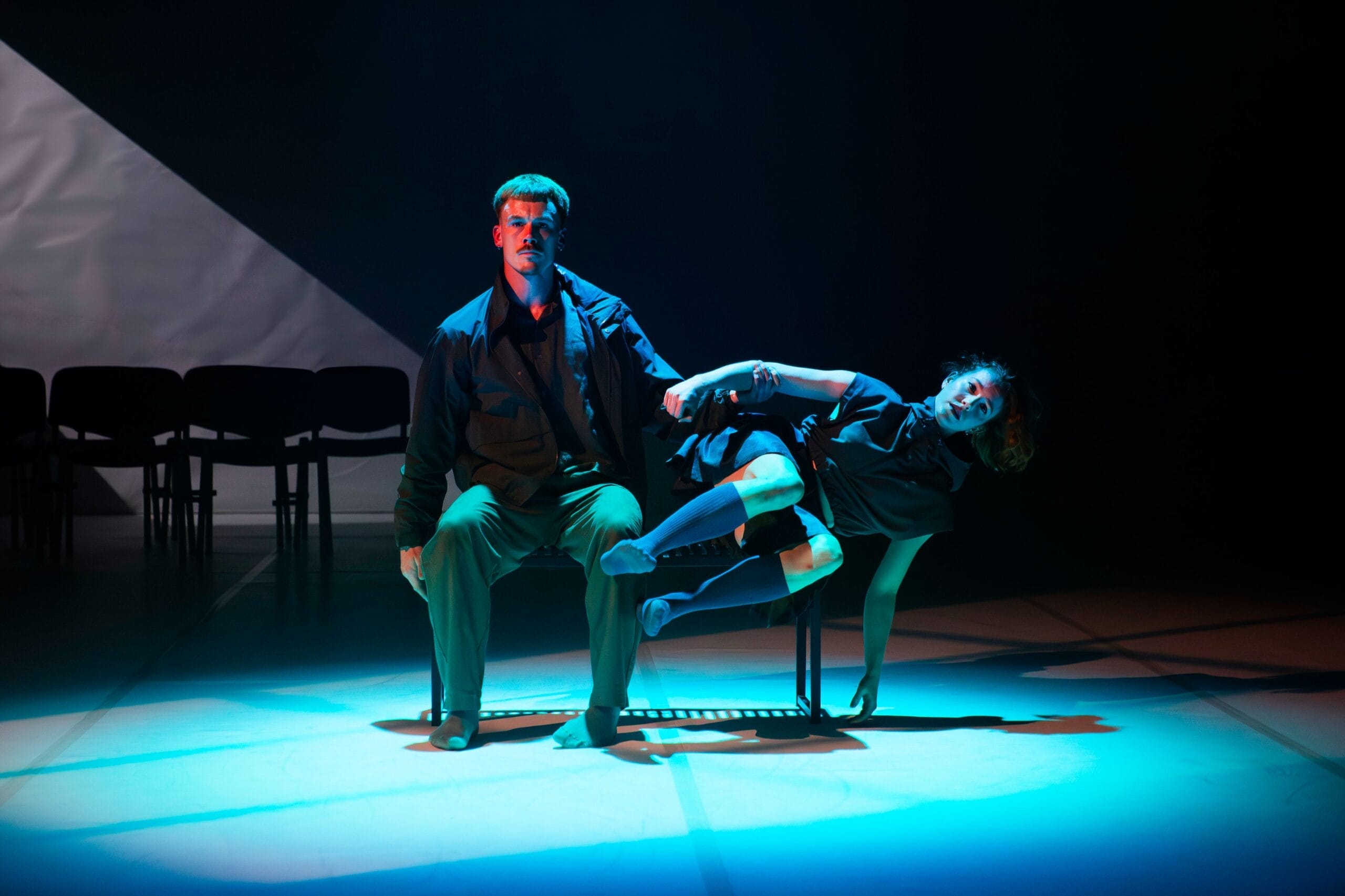 A man sits on a bench under stage lighting while a woman reclines across his lap, both barefoot and dressed in dark clothing on a dimly lit stage.