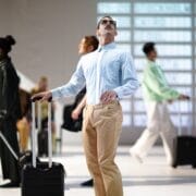 A man with a suitcase looks up while standing in front of a departure board, with other travelers walking by in an airport setting.