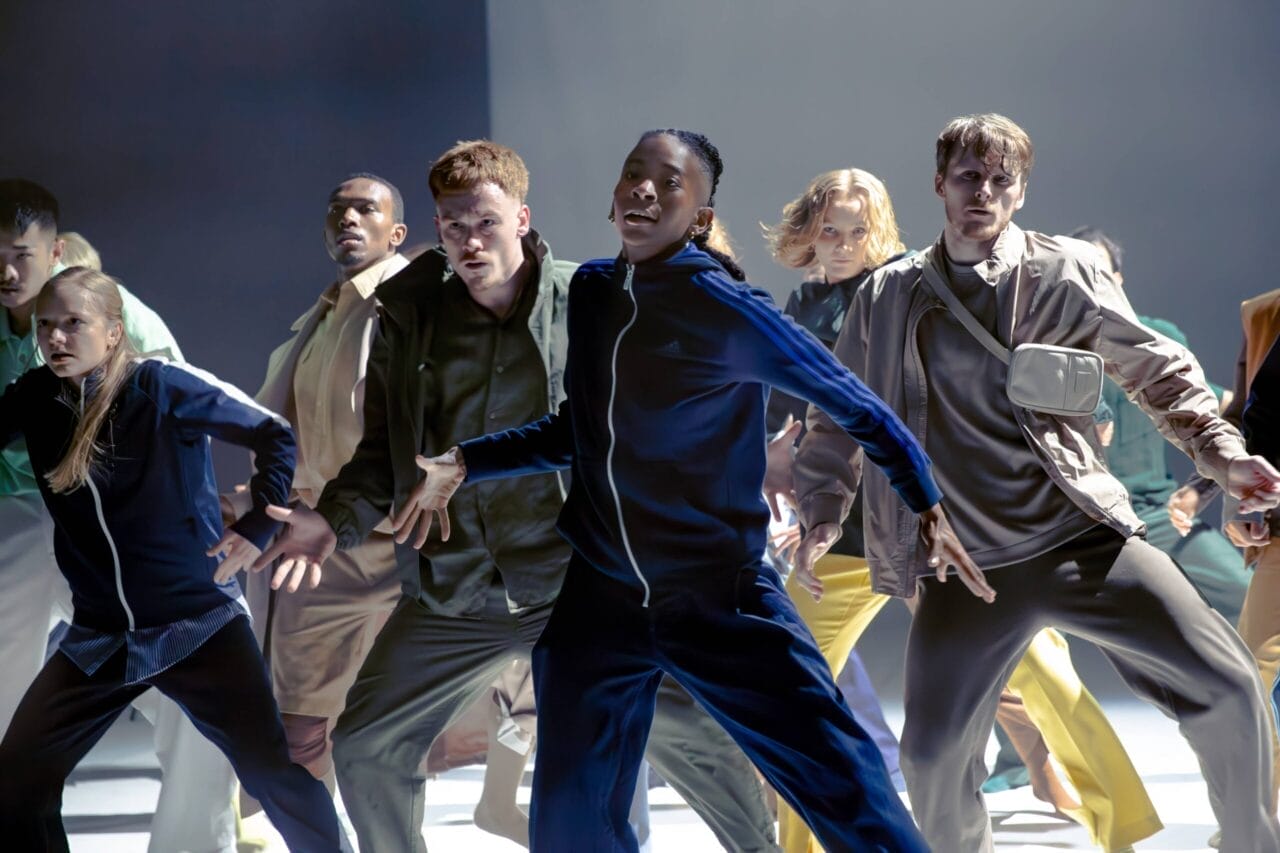 A group of people in casual clothing perform a synchronized dance on a brightly lit stage, with energetic and expressive poses.