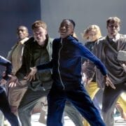 A group of people in casual clothing perform a synchronized dance on a brightly lit stage, with energetic and expressive poses.