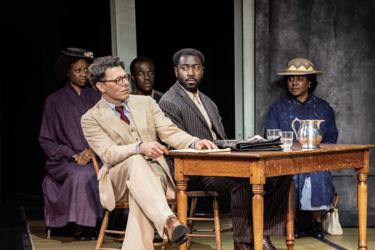 Five people in period clothing sit at and behind a wooden table on stage, evoking a courtroom scene reminiscent of Harper Lee's classic novel, To Kill a Mockingbird.