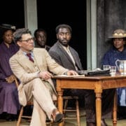 Five people in period clothing sit at and behind a wooden table on stage, evoking a courtroom scene reminiscent of Harper Lee's classic novel, To Kill a Mockingbird.