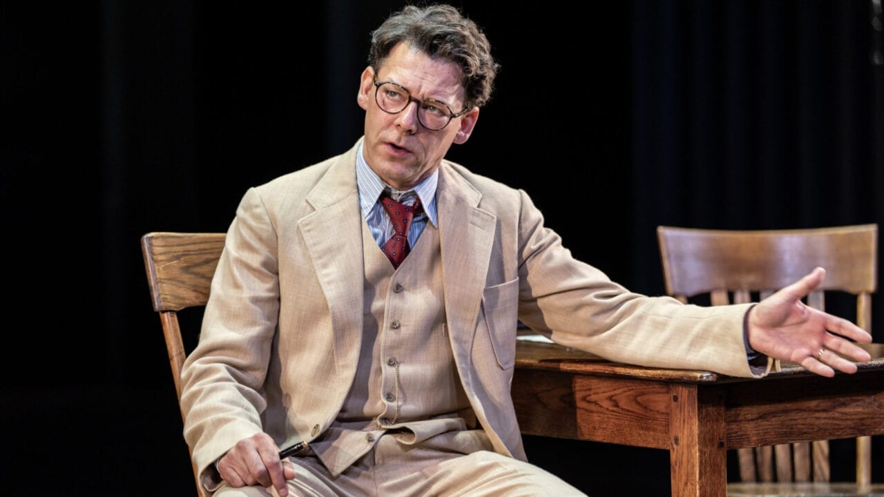A man in a beige suit and glasses sits at a wooden table, gesturing with one hand while speaking, reminiscent of Atticus Finch from Harper Lee's classic novel, To Kill a Mockingbird.