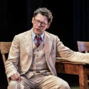 A man in a beige suit and glasses sits at a wooden table, gesturing with one hand while speaking, reminiscent of Atticus Finch from Harper Lee's classic novel, To Kill a Mockingbird.