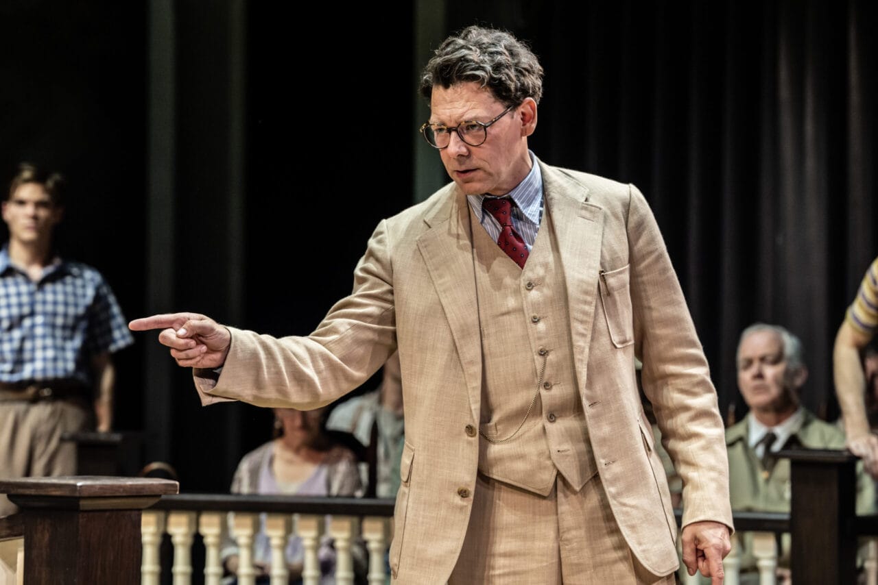 A man in a beige suit and glasses points his finger while speaking in a courtroom, evoking the iconic courtroom scenes from Harper Lee’s classic novel To Kill a Mockingbird, with people seated attentively in the background.