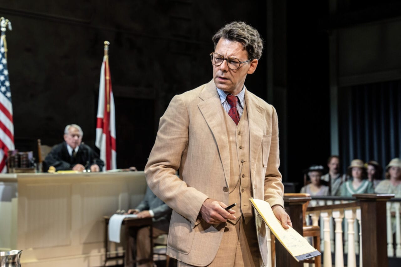 A man in a tan suit stands in a courtroom holding a yellow notepad, evoking scenes from Harper Lee’s classic novel To Kill a Mockingbird, with a judge, flags, and seated people in the background.