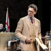 A man in a tan suit stands in a courtroom holding a yellow notepad, evoking scenes from Harper Lee’s classic novel To Kill a Mockingbird, with a judge, flags, and seated people in the background.