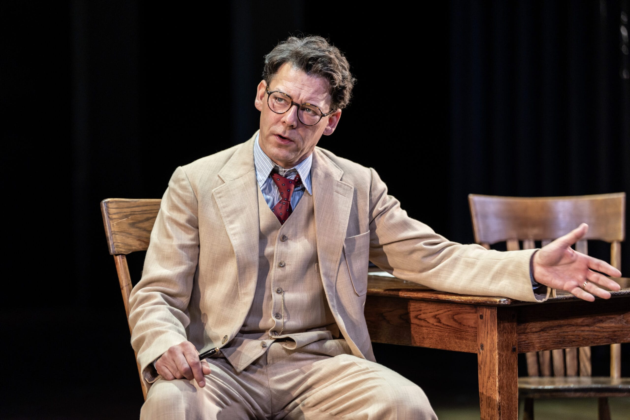 A man in a beige suit and glasses sits at a wooden table, gesturing with one hand while speaking, reminiscent of Atticus Finch from Harper Lee's classic novel, To Kill a Mockingbird.