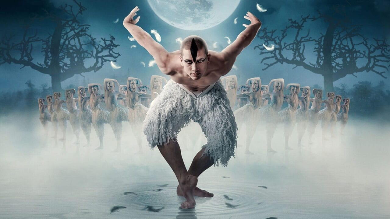 A male dancer dressed in white poses with arms raised, surrounded by dancers in a foggy, moonlit setting with leafless trees.