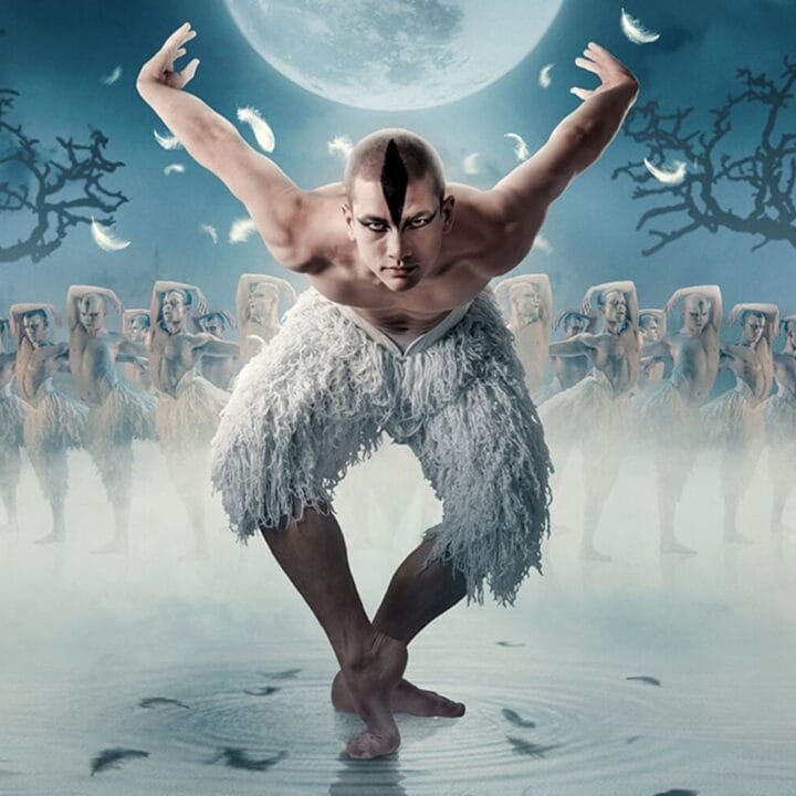A male dancer dressed in white poses with arms raised, surrounded by dancers in a foggy, moonlit setting with leafless trees.