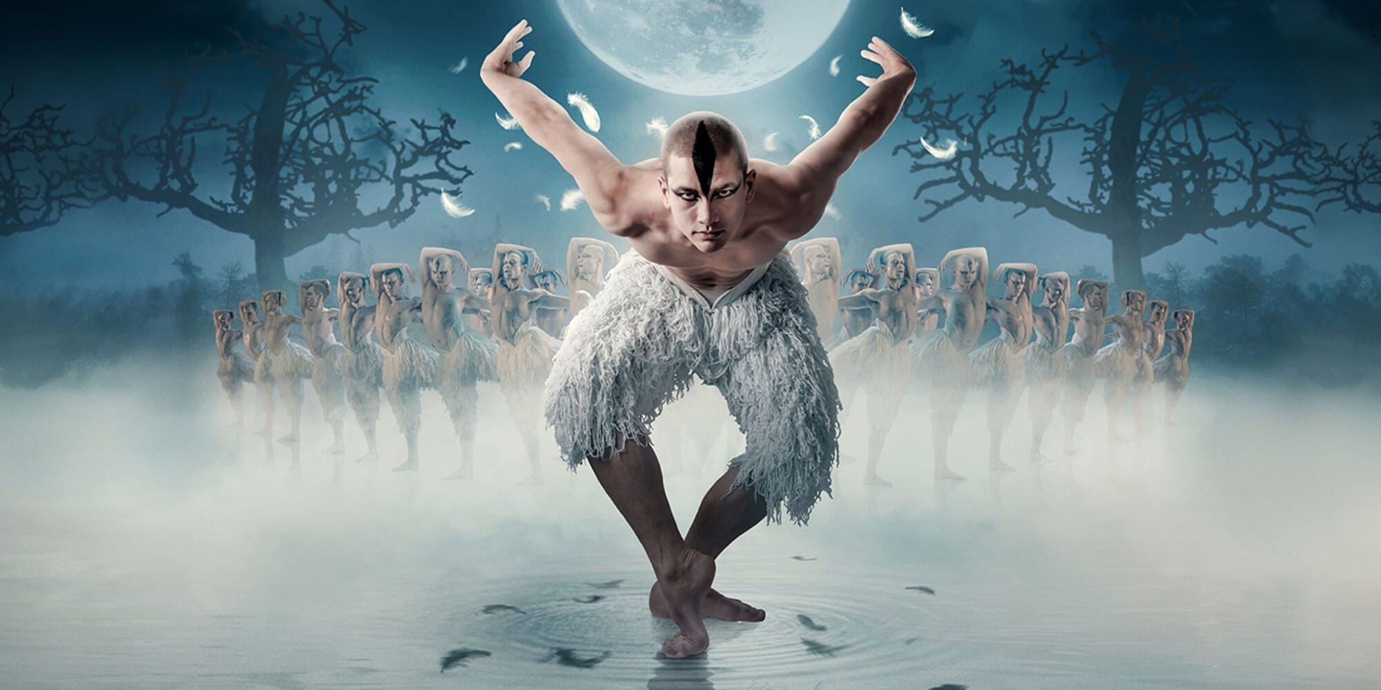A male dancer dressed in white poses with arms raised, surrounded by dancers in a foggy, moonlit setting with leafless trees.