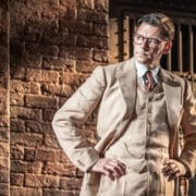 A man in a beige three-piece suit and glasses stands with hands on hips in front of a brick wall, evoking the timeless style of Harper Lee’s classic novel, To Kill a Mockingbird, as he looks thoughtfully to the side.