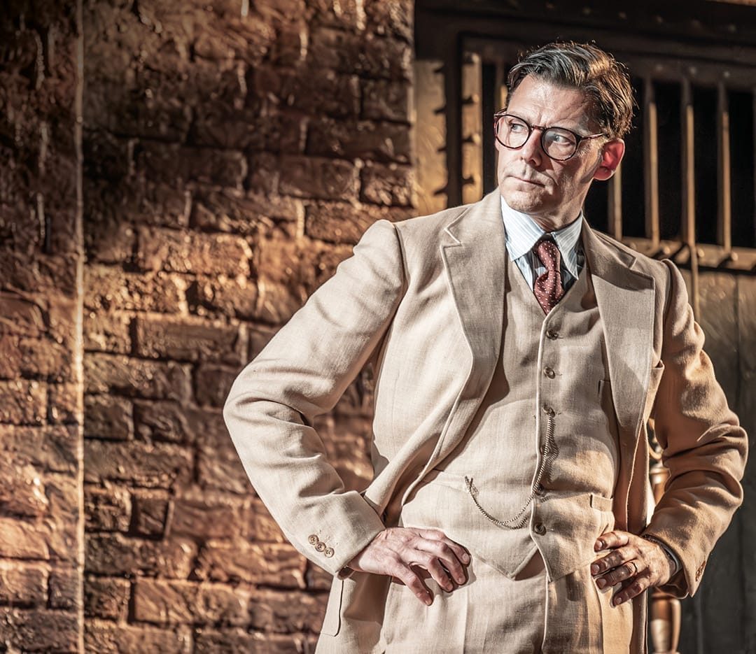 A man in a beige three-piece suit and glasses stands with hands on hips in front of a brick wall, evoking the timeless style of Harper Lee’s classic novel, To Kill a Mockingbird, as he looks thoughtfully to the side.