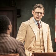 A man in a beige suit and glasses stands facing another seated man in a pinstripe suit, evoking a scene from Harper Lee’s classic novel, To Kill a Mockingbird, with an American flag visible in the background.