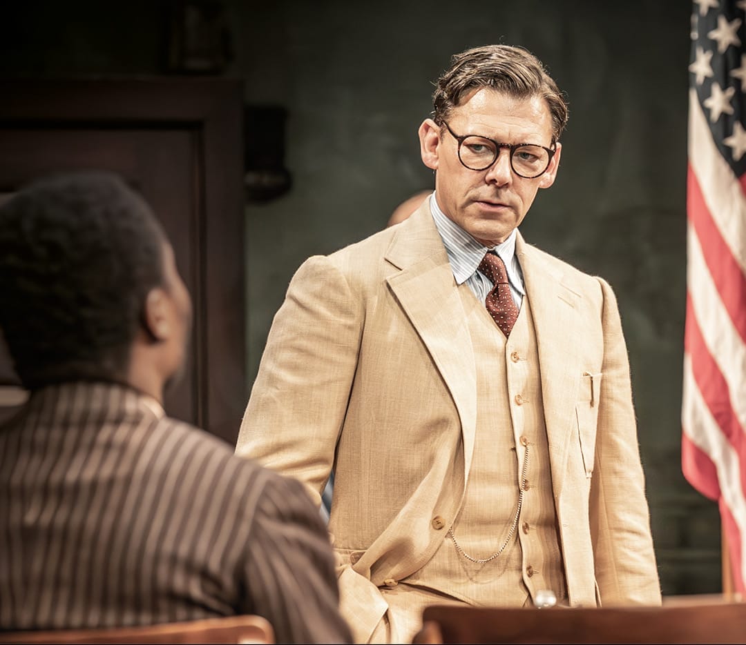 A man in a beige suit and glasses stands facing another seated man in a pinstripe suit, evoking a scene from Harper Lee’s classic novel, To Kill a Mockingbird, with an American flag visible in the background.