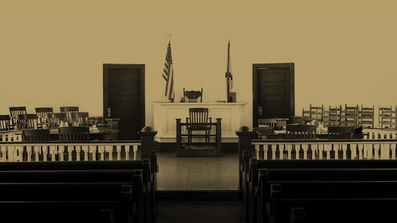 Empty courtroom with wooden benches, a judge’s bench, jury seats, and American and state flags in the background.