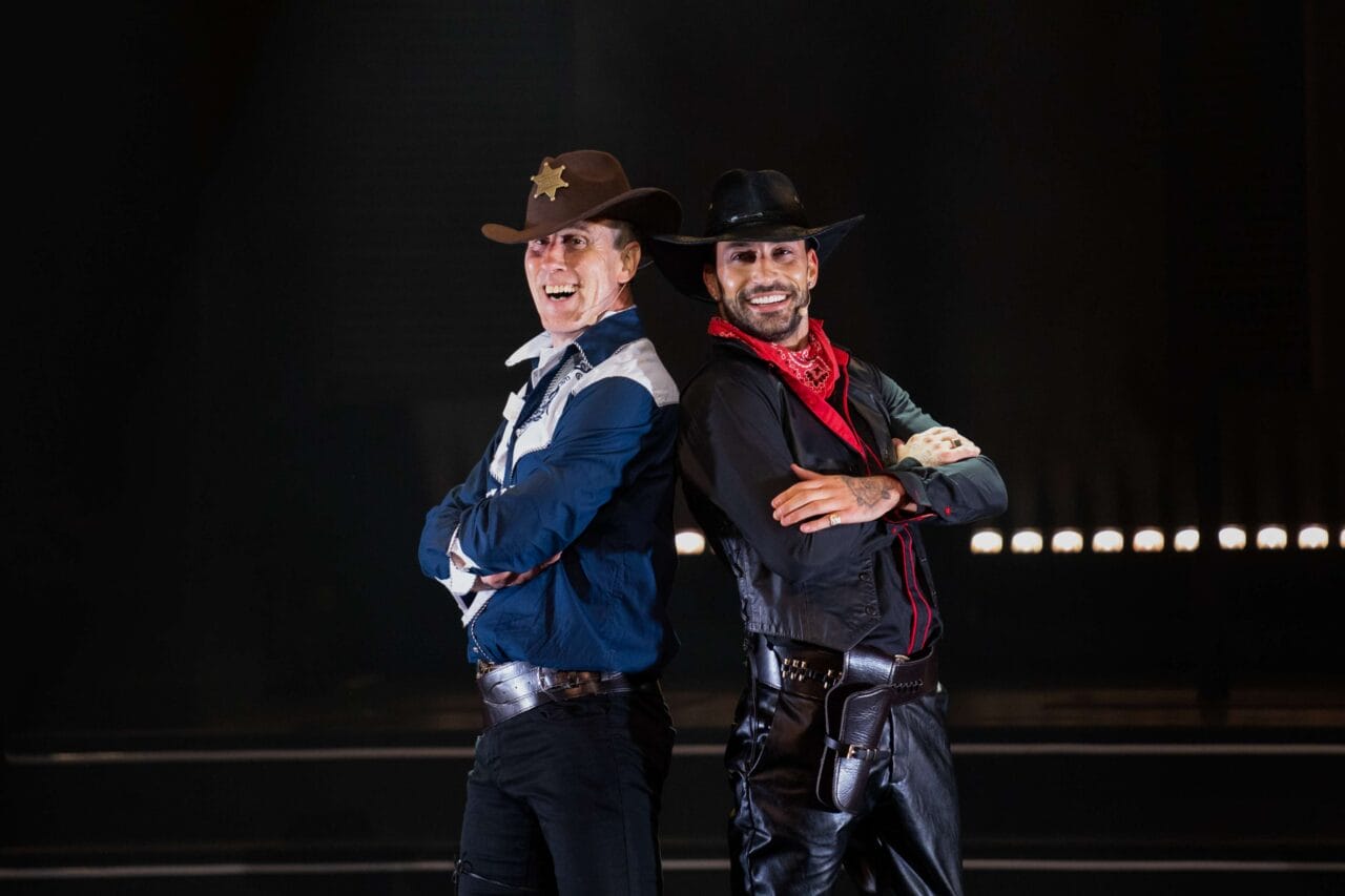 Two men dressed as cowboys, one in a sheriff's outfit with a badge and hat, the other in black with a red bandana, stand back to back and smile on stage.