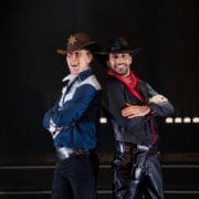 Two men dressed as cowboys, one in a sheriff's outfit with a badge and hat, the other in black with a red bandana, stand back to back and smile on stage.