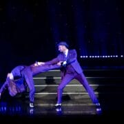 Two performers in suits and hats dance on stage; one bends backward with a hat in hand while the other supports him, under dramatic purple lighting.