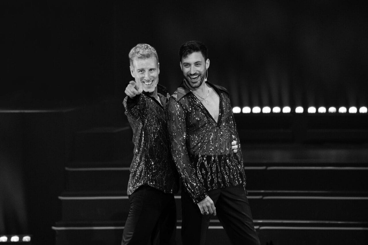 Two men wearing matching sparkling shirts stand on stage, smiling; one points towards the camera while the other has his arm around him. Steps and stage lights are visible in the background.