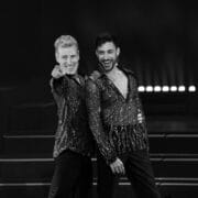 Two men wearing matching sparkling shirts stand on stage, smiling; one points towards the camera while the other has his arm around him. Steps and stage lights are visible in the background.