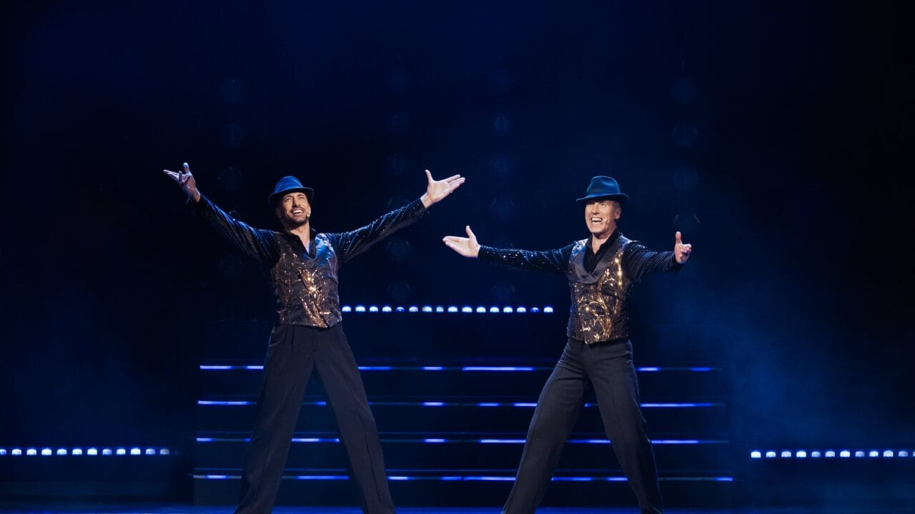 Two male performers wearing black hats, black trousers, and sequined vests stand onstage with arms outstretched, illuminated by stage lights against a dark background.