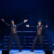 Two male performers wearing black hats, black trousers, and sequined vests stand onstage with arms outstretched, illuminated by stage lights against a dark background.