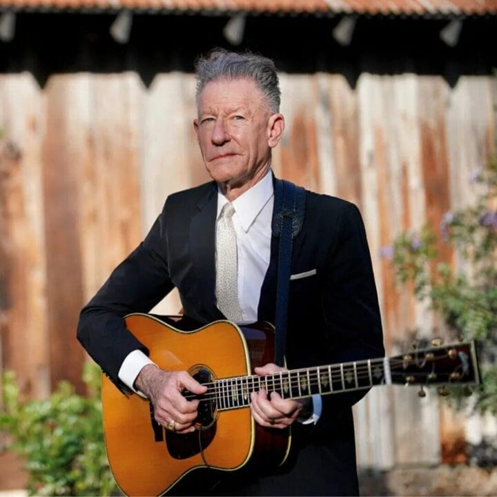 Lyle Lovett in a black suit and tie stands outdoors in front of a wooden wall, playing an acoustic guitar.