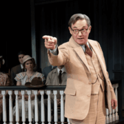 A man in a beige suit stands in front of a seated group, pointing forward, with a wooden railing and dark background behind him, suggesting a courtroom or theatrical scene.