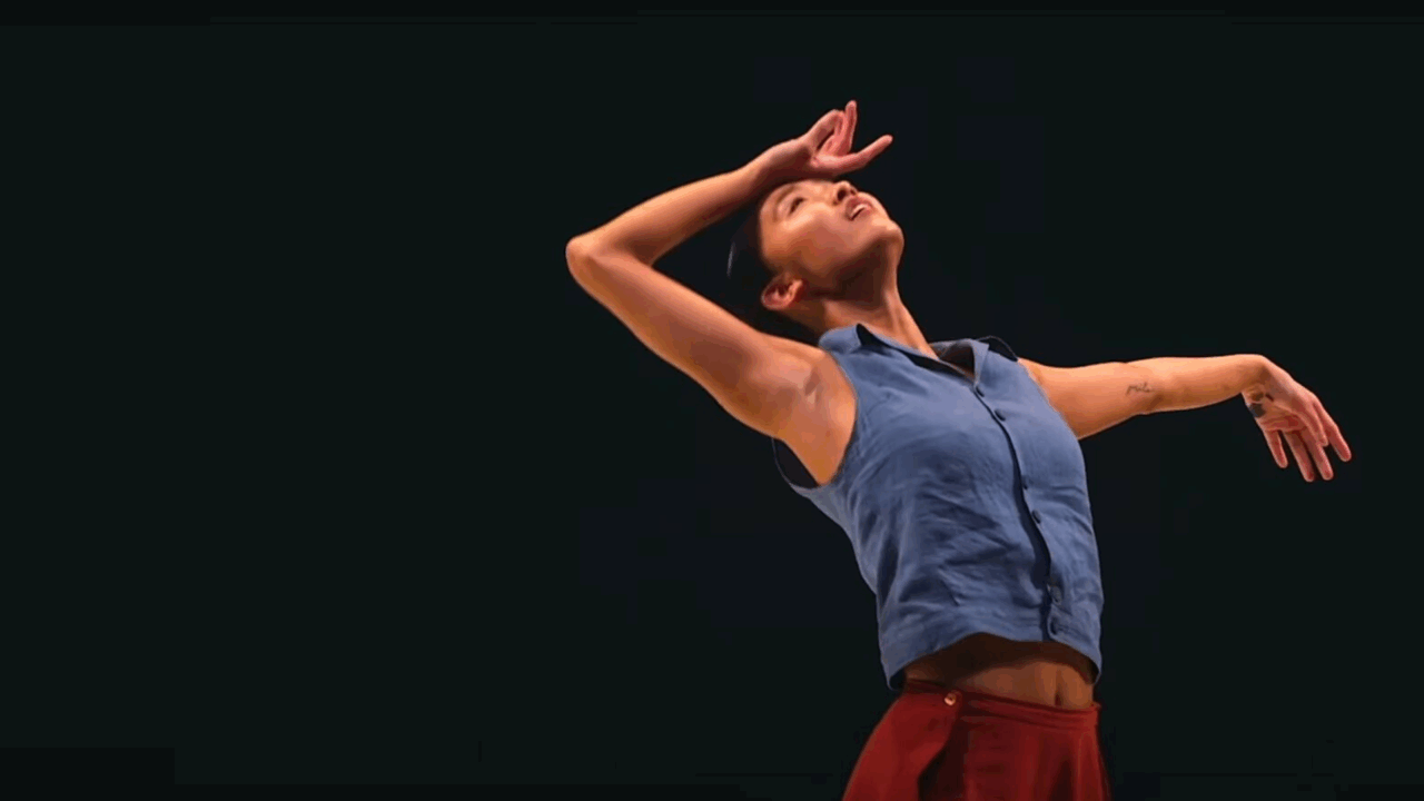 A person in a blue sleeveless shirt and red pants poses with one arm curved over their head and the other extended outward against a dark background.