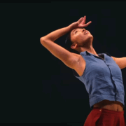 A person in a blue sleeveless shirt and red pants poses with one arm curved over their head and the other extended outward against a dark background.