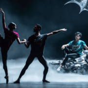 Three dancers perform on stage; two pose gracefully in the foreground, while one sits on a metallic sculpture resembling a car with a winged figure. Mist covers the floor.