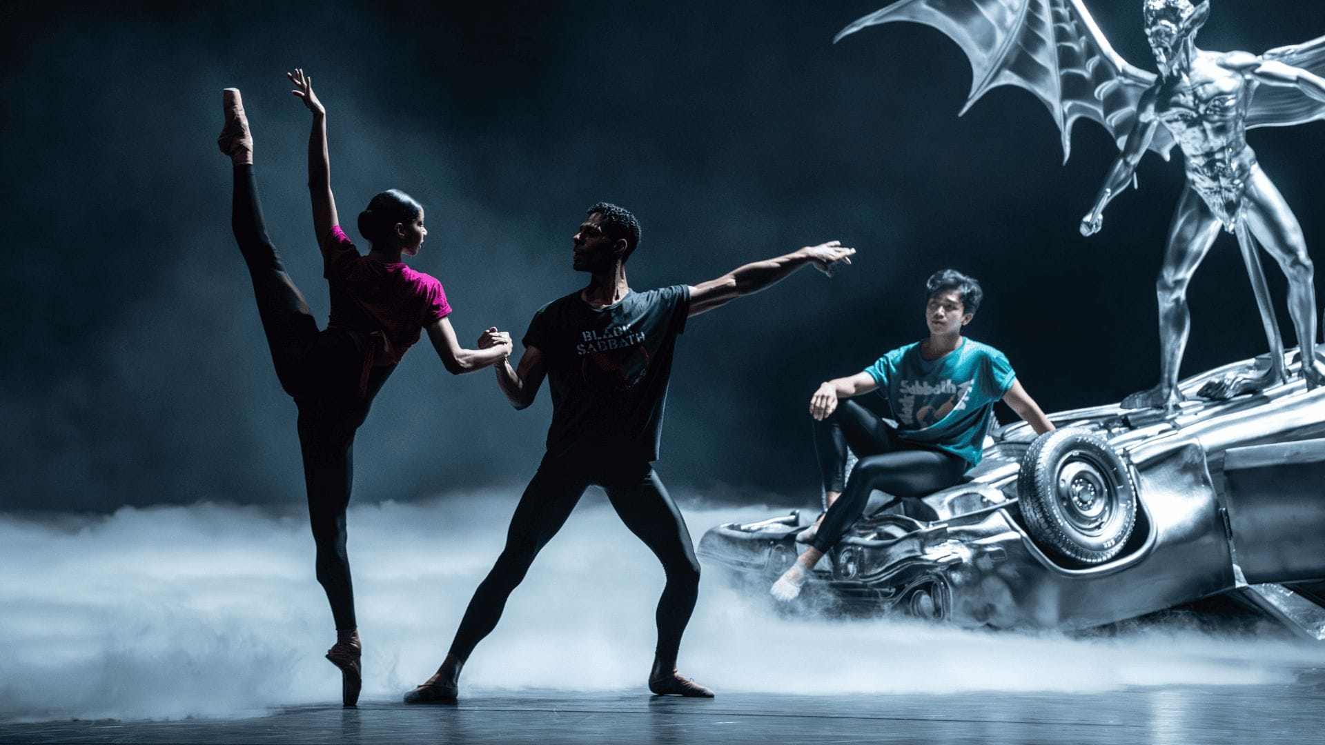 Three dancers perform on stage; two pose gracefully in the foreground, while one sits on a metallic sculpture resembling a car with a winged figure. Mist covers the floor.