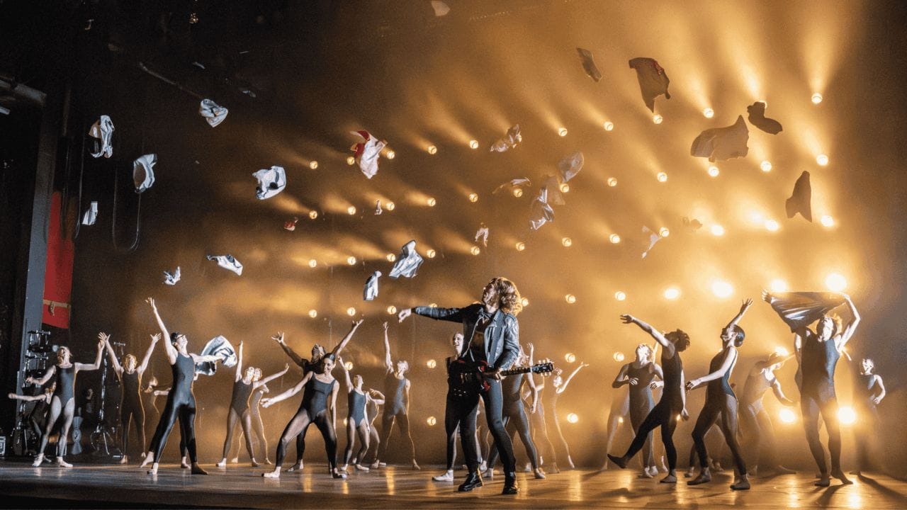 A group of dancers perform energetically on stage as clothes are thrown into the air, with dramatic lighting and a guitarist in the center.