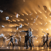 A group of dancers perform energetically on stage as clothes are thrown into the air, with dramatic lighting and a guitarist in the center.