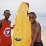 Two boys stand on a sandy beach, one in a red Captain America tank top, the other shirtless with a yellow surfboard, ready for adventure. The scene echoes the spirit of the Ocean Film Festival with waves and clouds in the background.