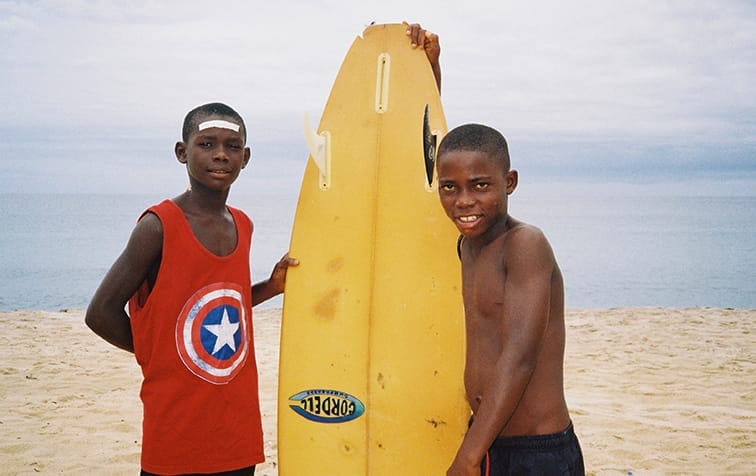 Two boys stand on a sandy beach, one in a red Captain America tank top, the other shirtless with a yellow surfboard, ready for adventure. The scene echoes the spirit of the Ocean Film Festival with waves and clouds in the background.