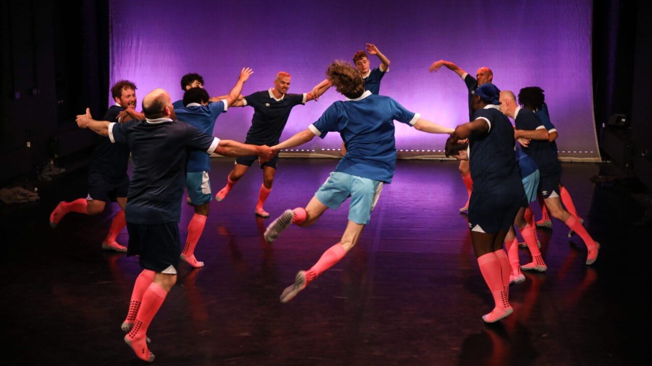 A group of people in blue shirts and pink knee socks hold hands in a circle, jumping in unison on a stage with a purple-lit backdrop.