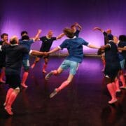 A group of people in blue shirts and pink knee socks hold hands in a circle, jumping in unison on a stage with a purple-lit backdrop.