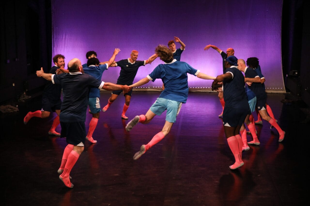 A group of people in matching blue shirts and pink knee-high socks jump and dance in a circle on a stage with a purple-lit background.